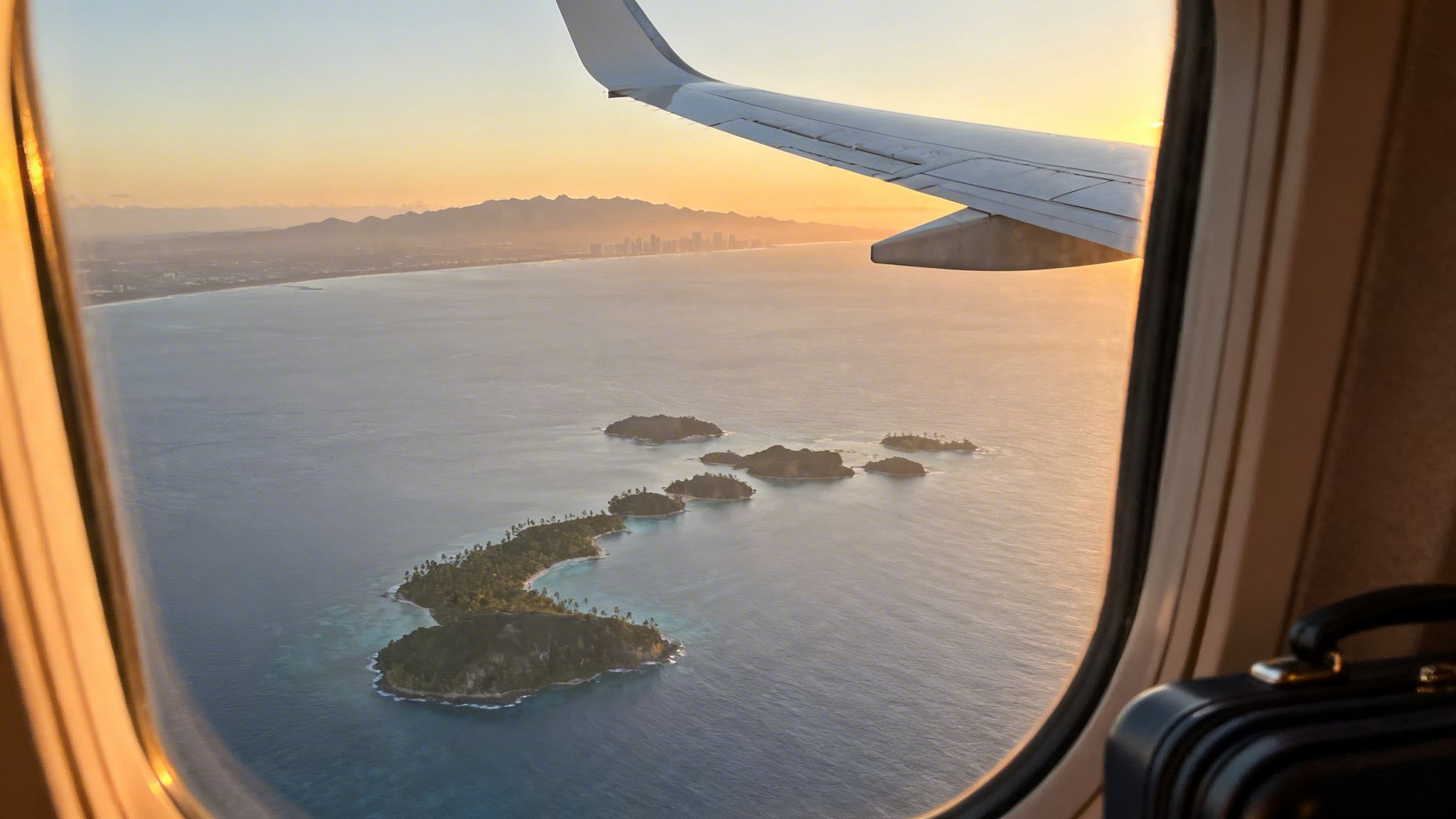 Aerial view from an airplane window showing tropical islands, a distant city, and mountains at sunrise.