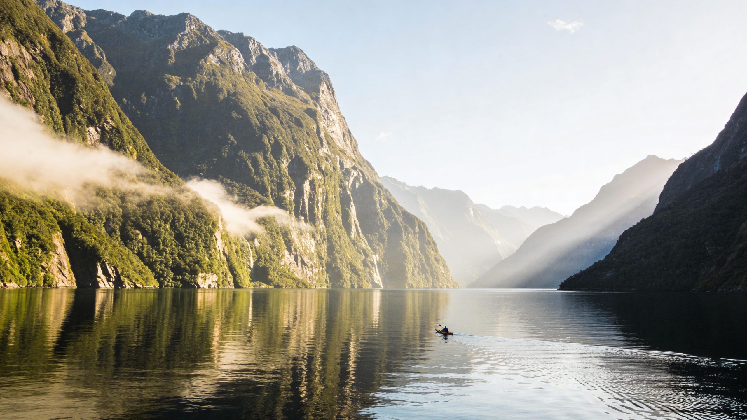 A person kayaking on calm water surrounded by majestic mountains and mist in Milford Sound, New Zealand.